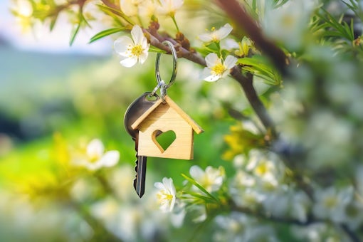 On a branch speckled with flowers, a key dangles, hinting at the joys of homeownership and new starts. A key ring with a house shape hangs on a blooming branch, embodying home dreams.