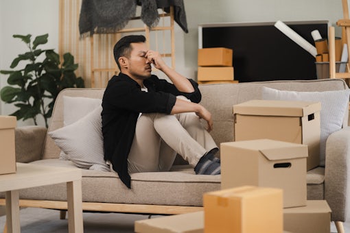 Exhausted Male sit rest on sofa in living room near heap of cardboard boxes feel unmotivated to unpack their belongings. Financial problem, debt and eviction, house debt bankrupt stress concept.