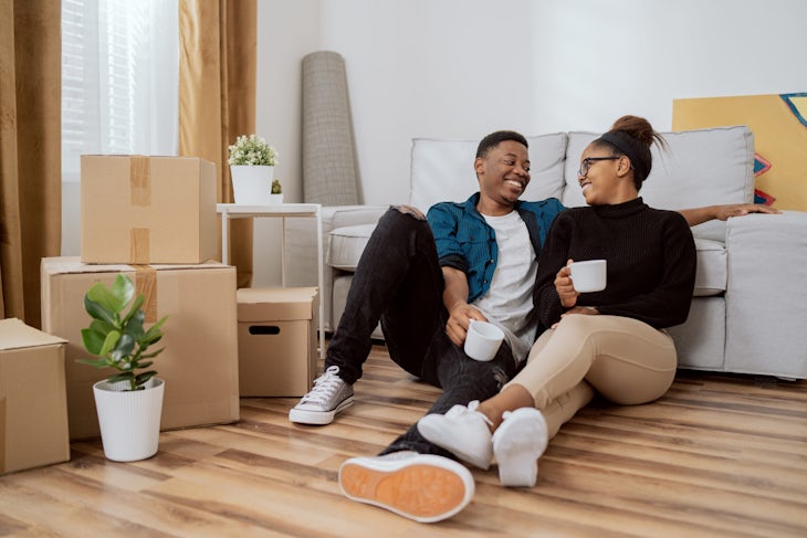Smiling couple is sitting on floor relaxing after moving in, drinking coffee in their new apartment, around them boxes with unpacked things, husband embraces his wife and looks at each other happily