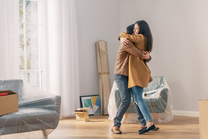 Happy and Excited Young Couple Look Around In Wonder at their Newly Purchased / Rented Apartment. Beautiful People Happily Embracing. Big Bright Modern Home with Cardboard Boxes Ready to Unpack.