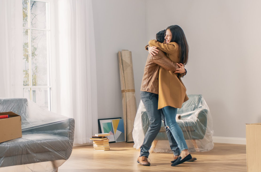 Happy and Excited Young Couple Look Around In Wonder at their Newly Purchased / Rented Apartment. Beautiful People Happily Embracing. Big Bright Modern Home with Cardboard Boxes Ready to Unpack.