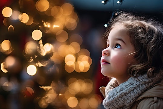 Young cute girl in awe of twinkling lights and ornaments on majestic Christmas tree. Sense of wonder and joy.